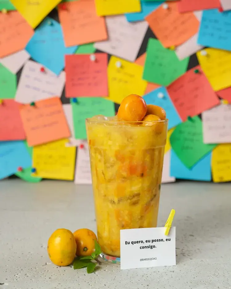 Um copo grande de bebida tropical com frutas, em frente a um fundo colorido de papéis adesivos. O copo contém uma mistura de frutas e é decorado com frutas em cima. A bebida simboliza um ambiente festivo e de celebração no Bar Seu João.