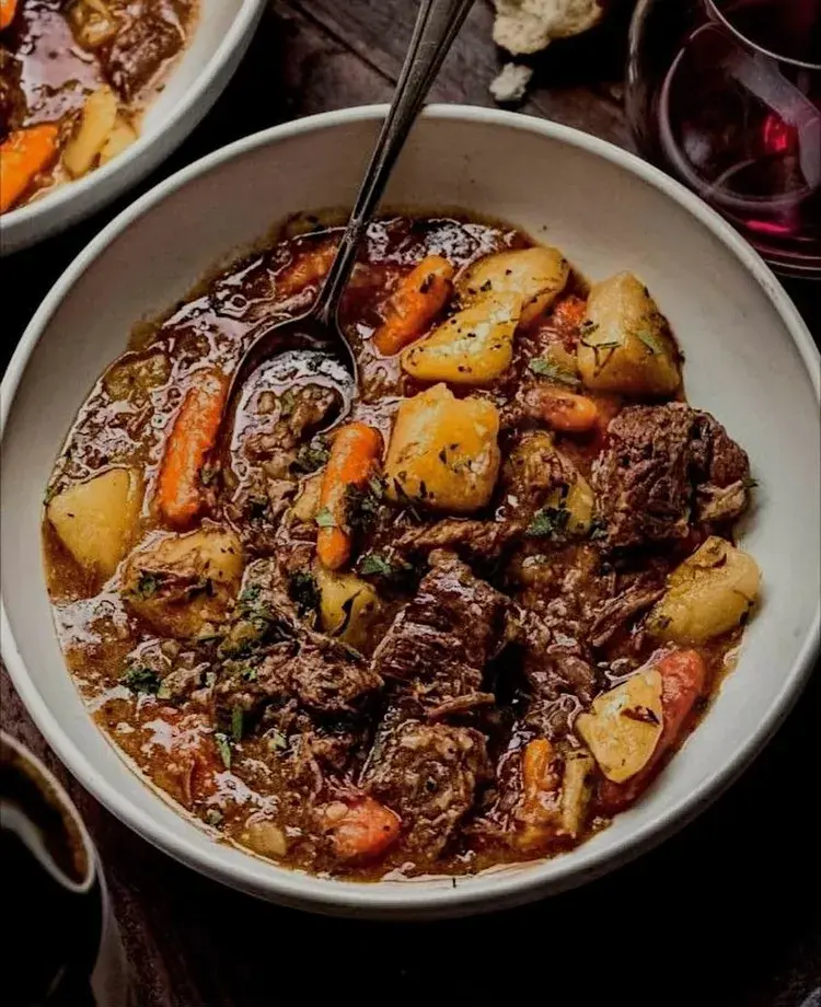 Prato de Cozido Português, servido em uma tigela branca, com carne, cenouras e batatas, em um caldo saboroso. A imagem mostra uma colher no prato e um copo de vinho ao fundo.