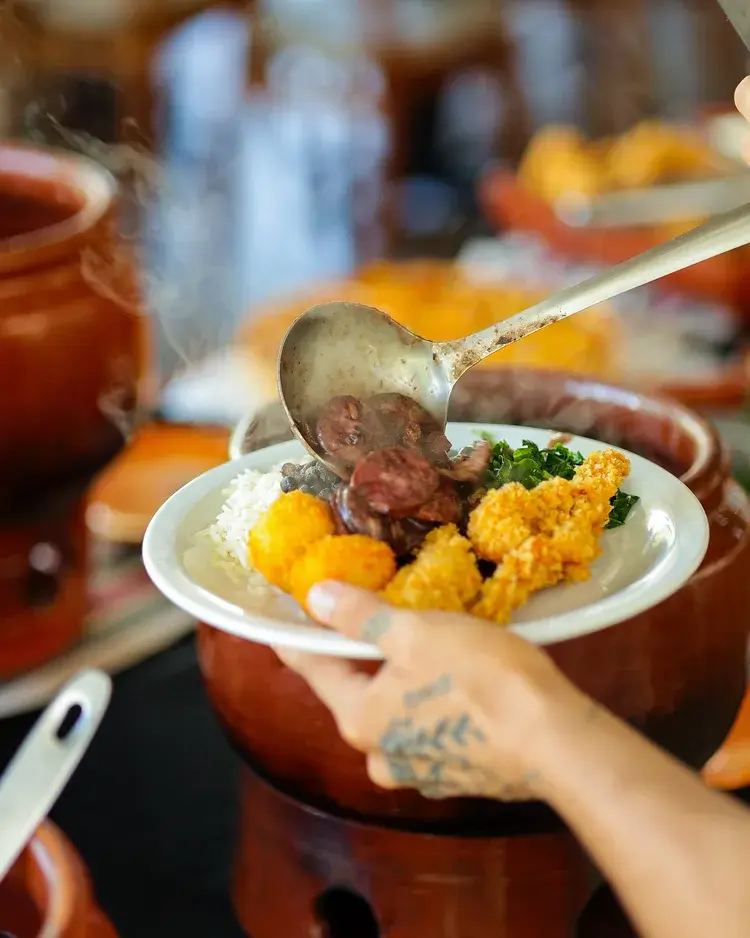 Uma pessoa servindo feijoada em um prato, com arroz e acompanhamentos, em um ambiente acolhedor no Johns Grill.