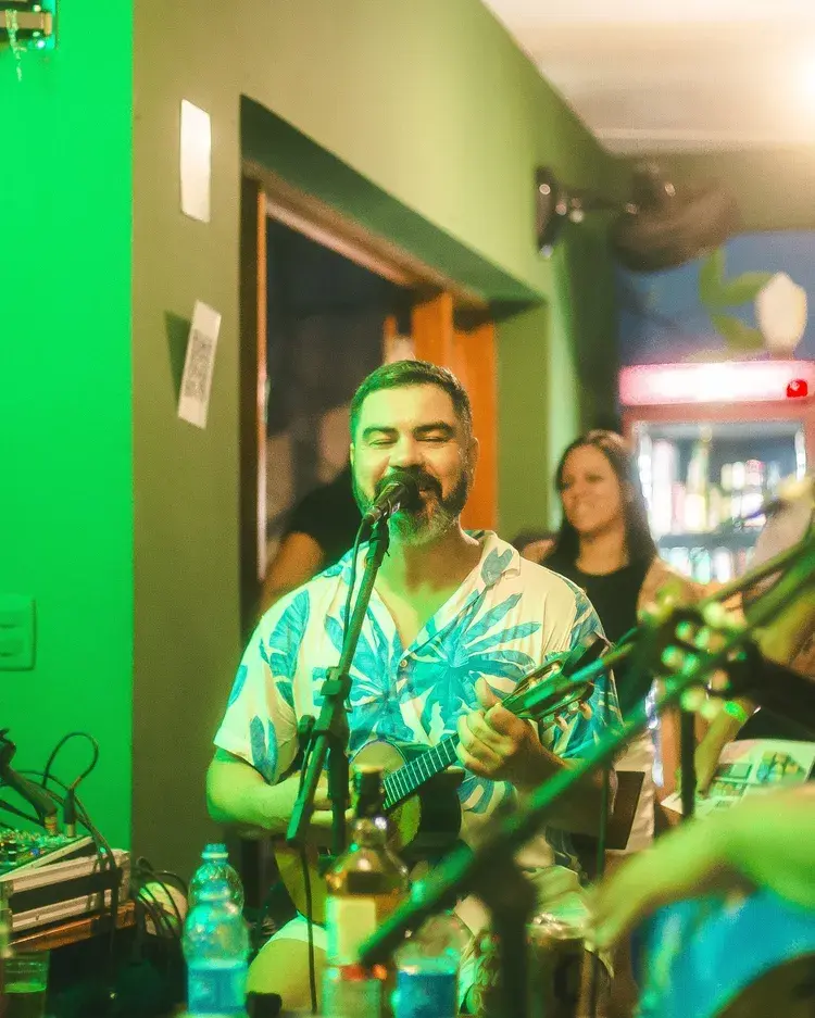 Imagem de um cantor sorrindo enquanto toca violão em um ambiente iluminado de verde, com pessoas ao fundo. O evento acontece no Recanto Jampa, em João Pessoa.