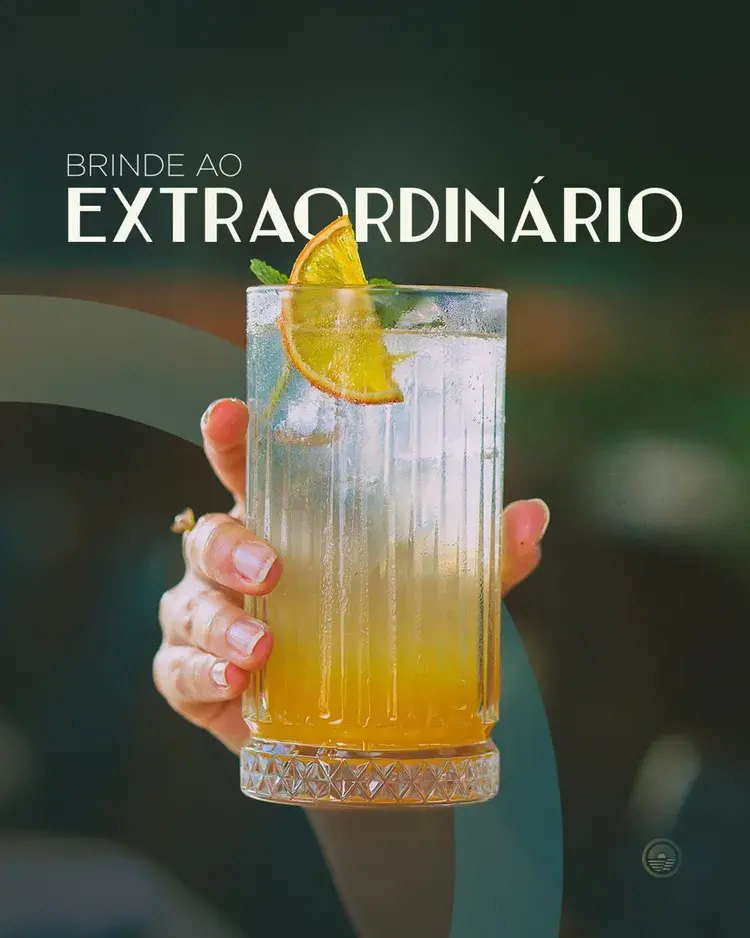 Imagem de um drink sofisticado sendo segurado por uma mão, com um fundo desfocado. O drink é decorado com uma fatia de laranja e folhas de hortelã, destacando a sofisticação do ambiente.