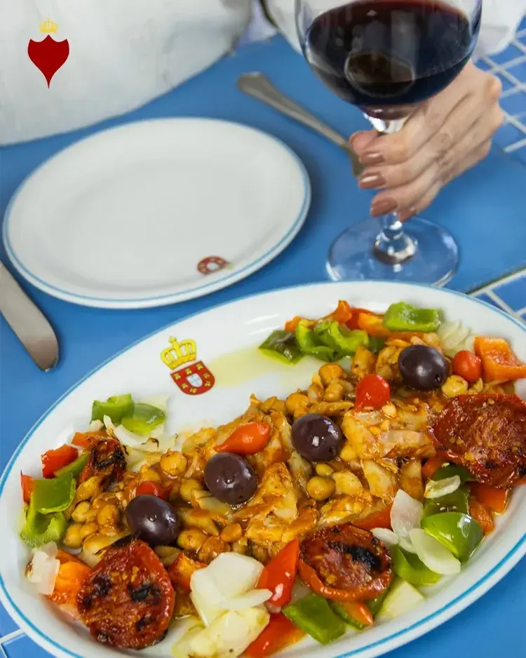 Prato de Bacalhau dos Passos servido em uma bandeja, com legumes coloridos e a marca da Casa do Bacalhau ao fundo.