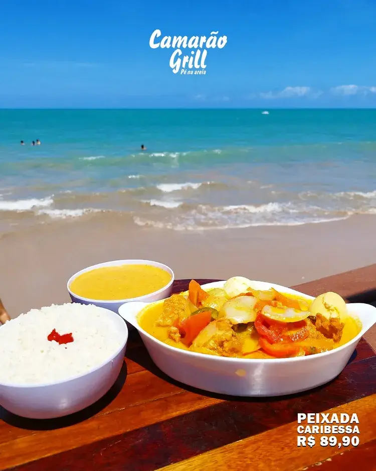 Imagem do Camarão Grill com um prato de caranguejo, cerveja Itaipava e uma bebida tropical, com o mar ao fundo.