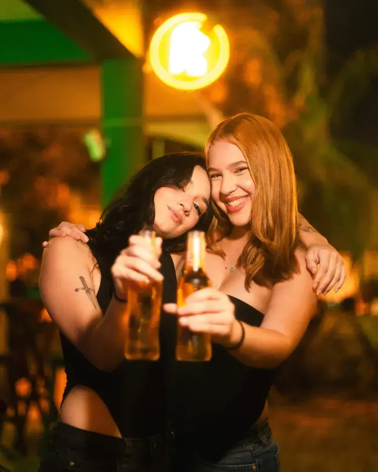 Duas mulheres sorrindo e segurando bebidas, celebrando em um ambiente festivo, com a iluminação do Recanto Jampa ao fundo.