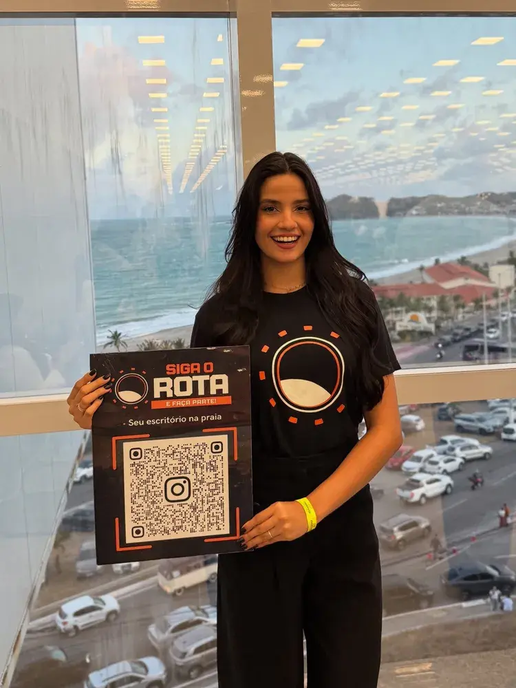 Mulher sorridente segurando um cartaz com QR code do Instagram e texto 'SIGA O ROTA E FAÇA PARTE! Seu escritório na praia', com vista da praia ao fundo, indicando presença do Rota 360 no Fórum de Negócios.