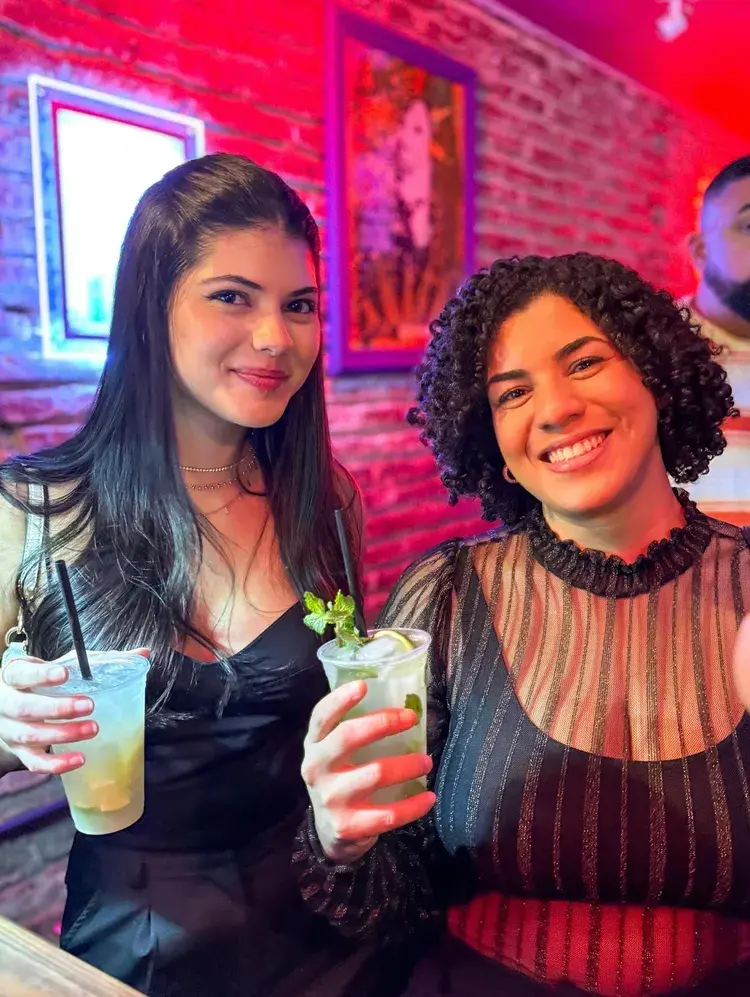 Duas mulheres sorrindo segurando drinks em copos plásticos, ambiente com iluminação vermelha e parede de tijolos ao fundo, texto na legenda menciona evento no bar Lili a partir das 18h.