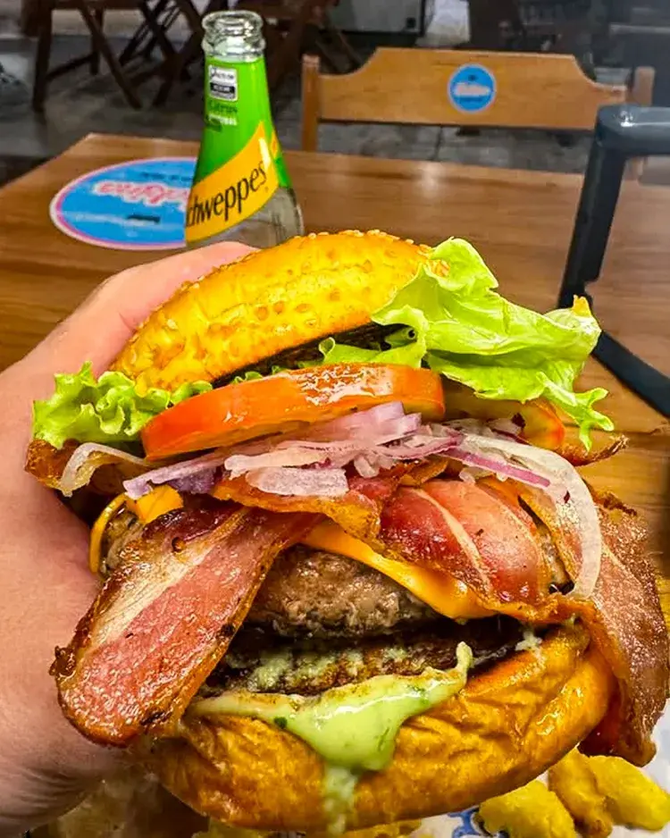 Imagem de um hambúrguer grande, chamado Shekiná Bacon, com bacon crocante, queijo derretido, carne suculenta e vegetais. Há uma garrafinha de refrigerante ao fundo, em um ambiente de restaurante.