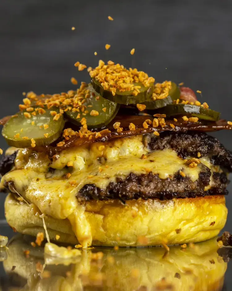 Imagem de um GB Cream do Gio Burger, mostrando dois smashes suculentos, quatro queijos, picles, bacon caramelizado e alho frito sobre um pão brioche.