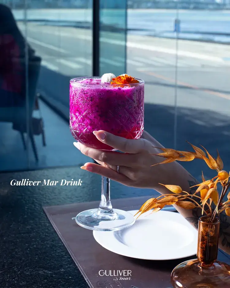 Imagem do Gulliver Mar Drink em uma taça alta, servida em um ambiente com vista para o mar. O drink é de cor vibrante e decorado com flores. O fundo mostra o pôr do sol na praia.