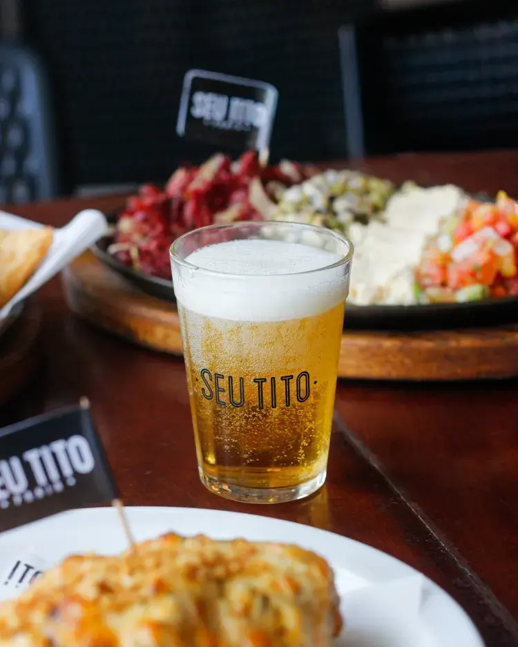 Copo de cerveja com a marca 'Seu Tito' e petiscos em uma mesa no bar Seu Tito. Ambiente animado e festivo.