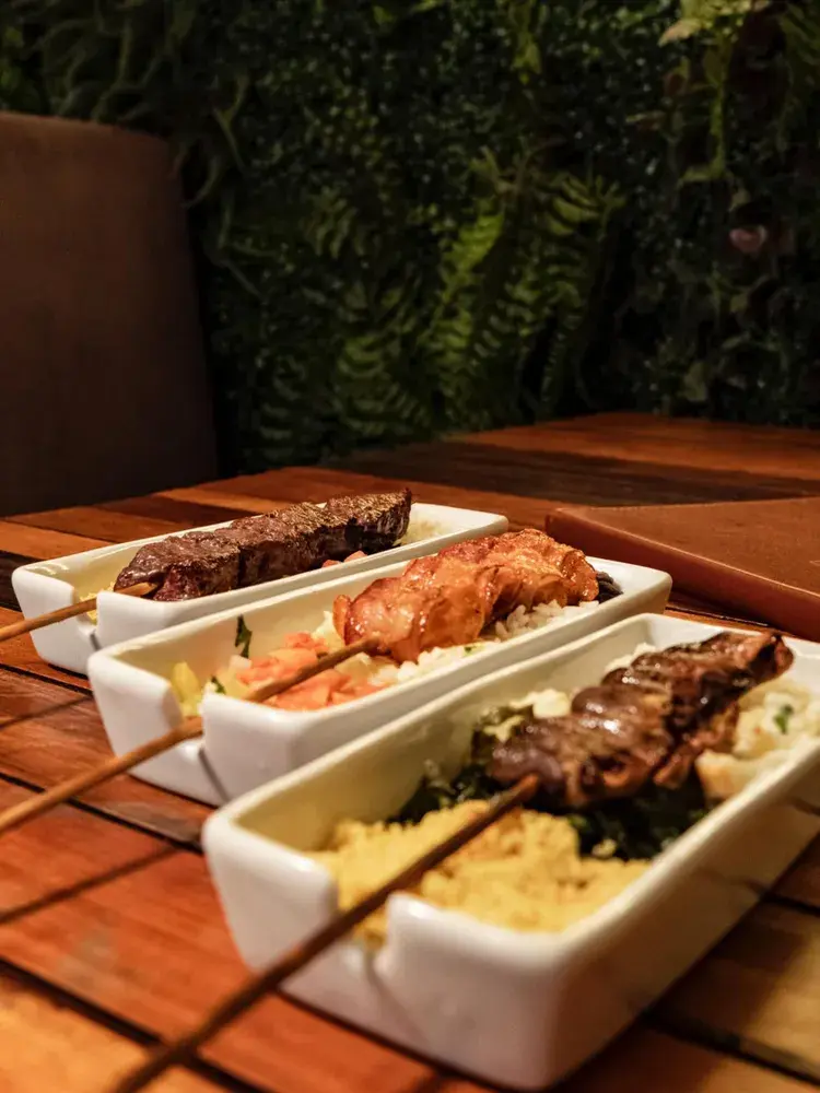 Imagem de espeto suculento e petiscos da Feijoada Manaíra, com ambiente acolhedor ao fundo.