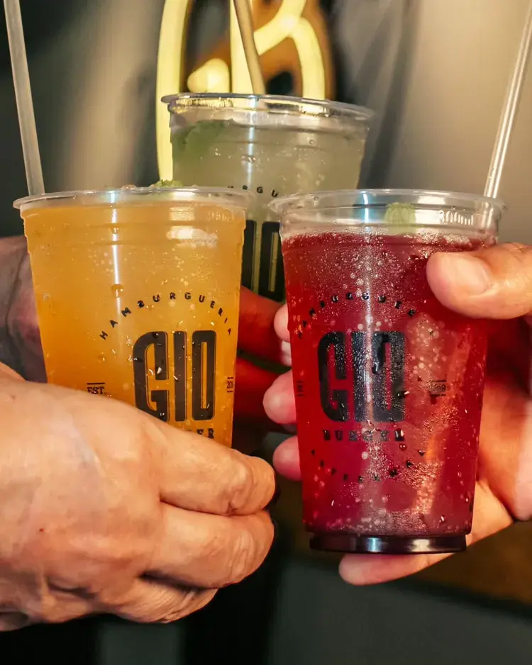 Três copos de sodas italianas do Gio Burger em mãos, com sabor de morango, maçã verde e maracujá. As sodas estão em copos transparentes com o logo do restaurante. Lugar: Avenida Acre 109, Bairro dos Estados, João Pessoa.