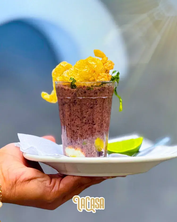 Um copo de caldinho repaginado sobre um prato branco, segurado por uma mão. O copo está decorado com crocante de batata e guarnição verde, com limão ao lado. A localização é La Casa, em Casa Forte, Recife.
