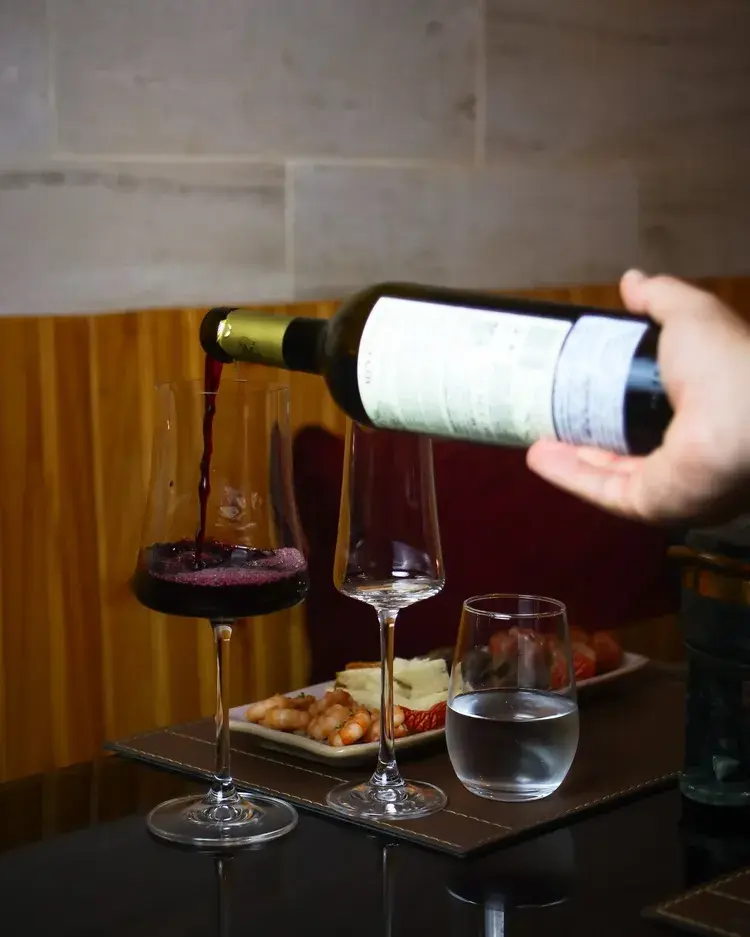 Um garçom serve vinho tinto em uma taça em uma mesa com pratos de camarão e frutas. A imagem mostra um ambiente acolhedor e preparado para receber os clientes.